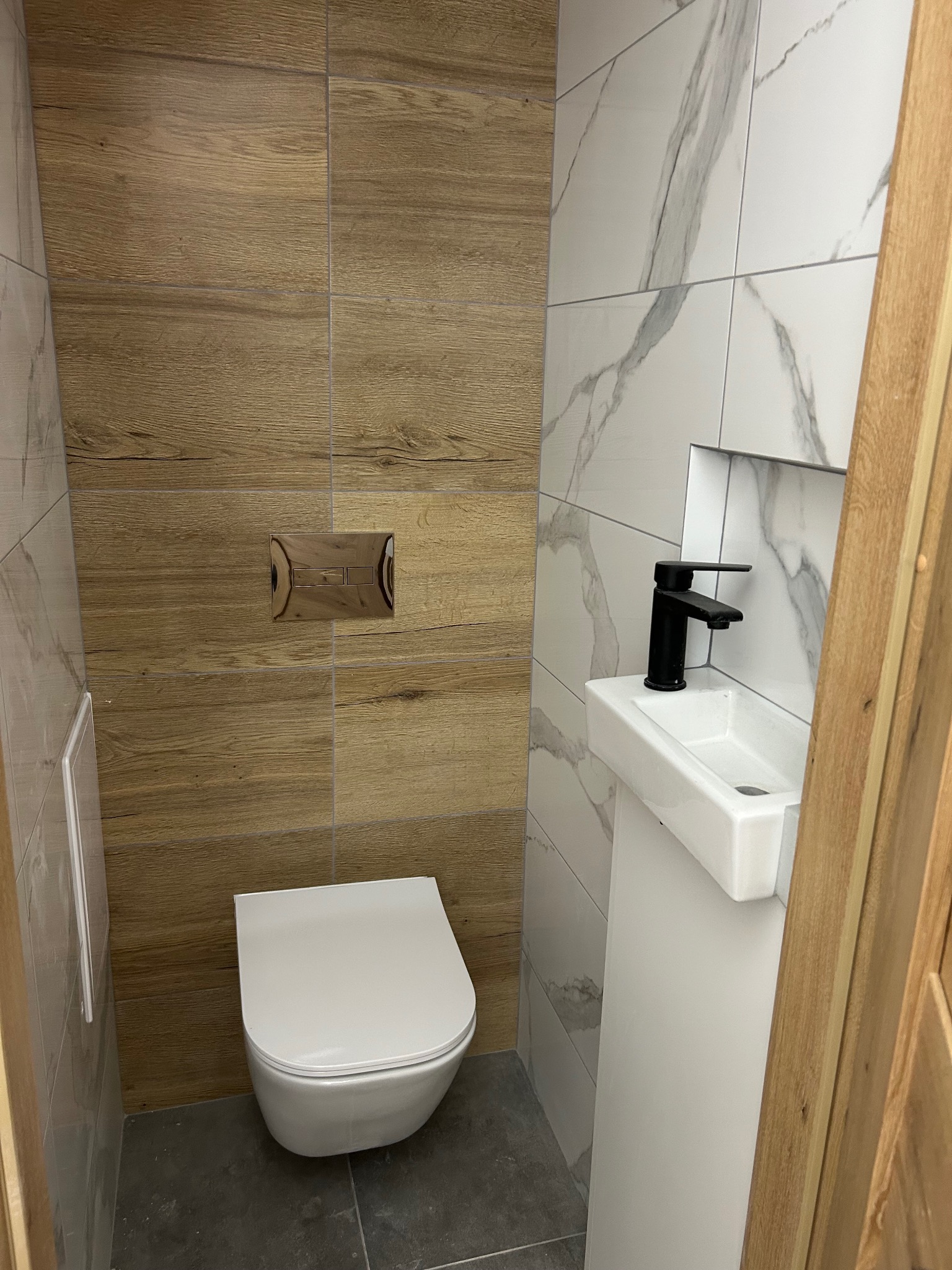Nowoczesna toaleta z drewnopodobnymi i marmurowymi płytkami, podwieszana miska WC i umywalka na postumencie z czarną armaturą, szara podłoga.