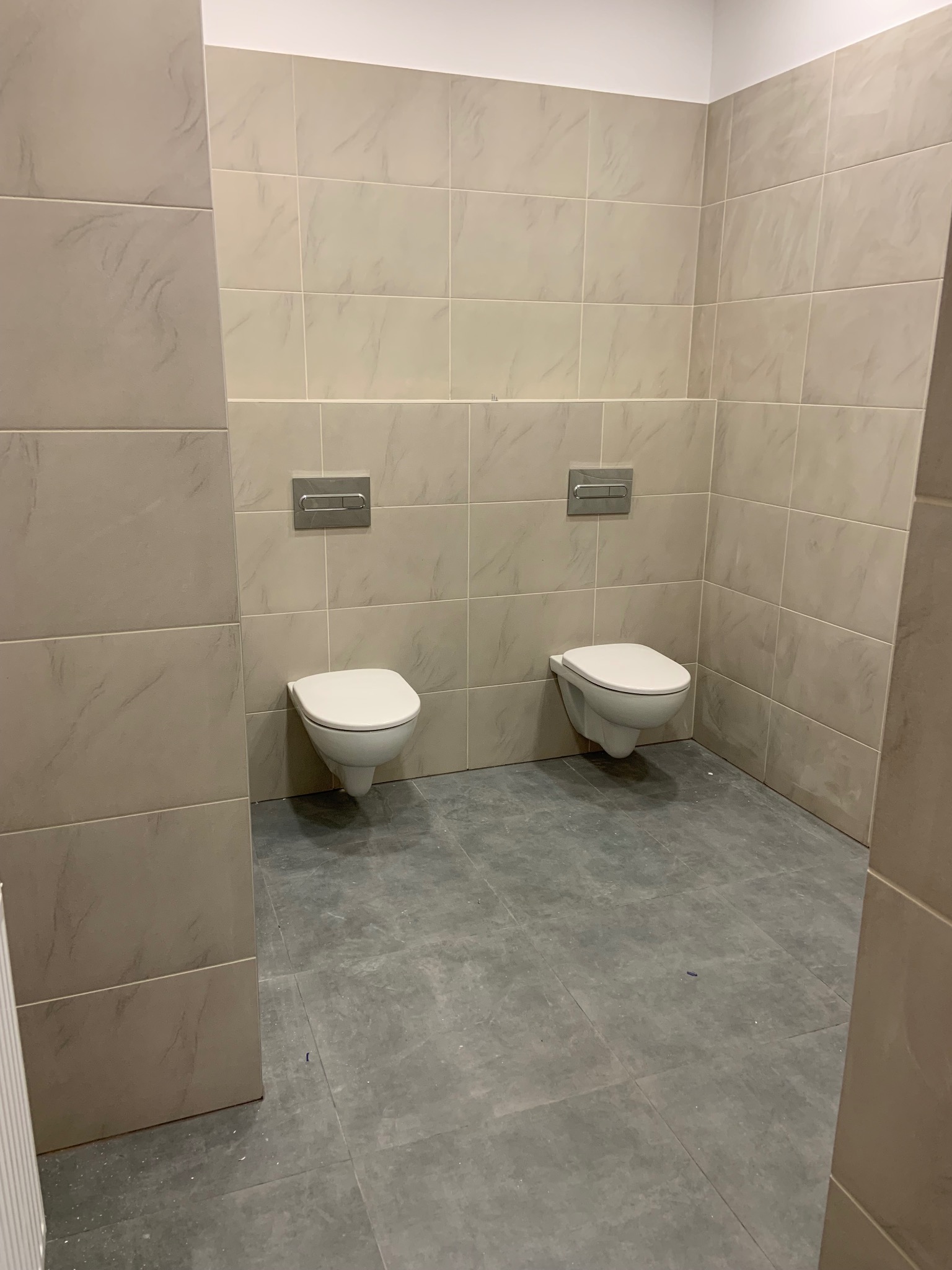 Wykończona łazienka z dwoma podwieszanymi toaletami i beżowymi płytkami. Szare płytki podłogowe. Widok na fragment wnętrza łazienki po remoncie.