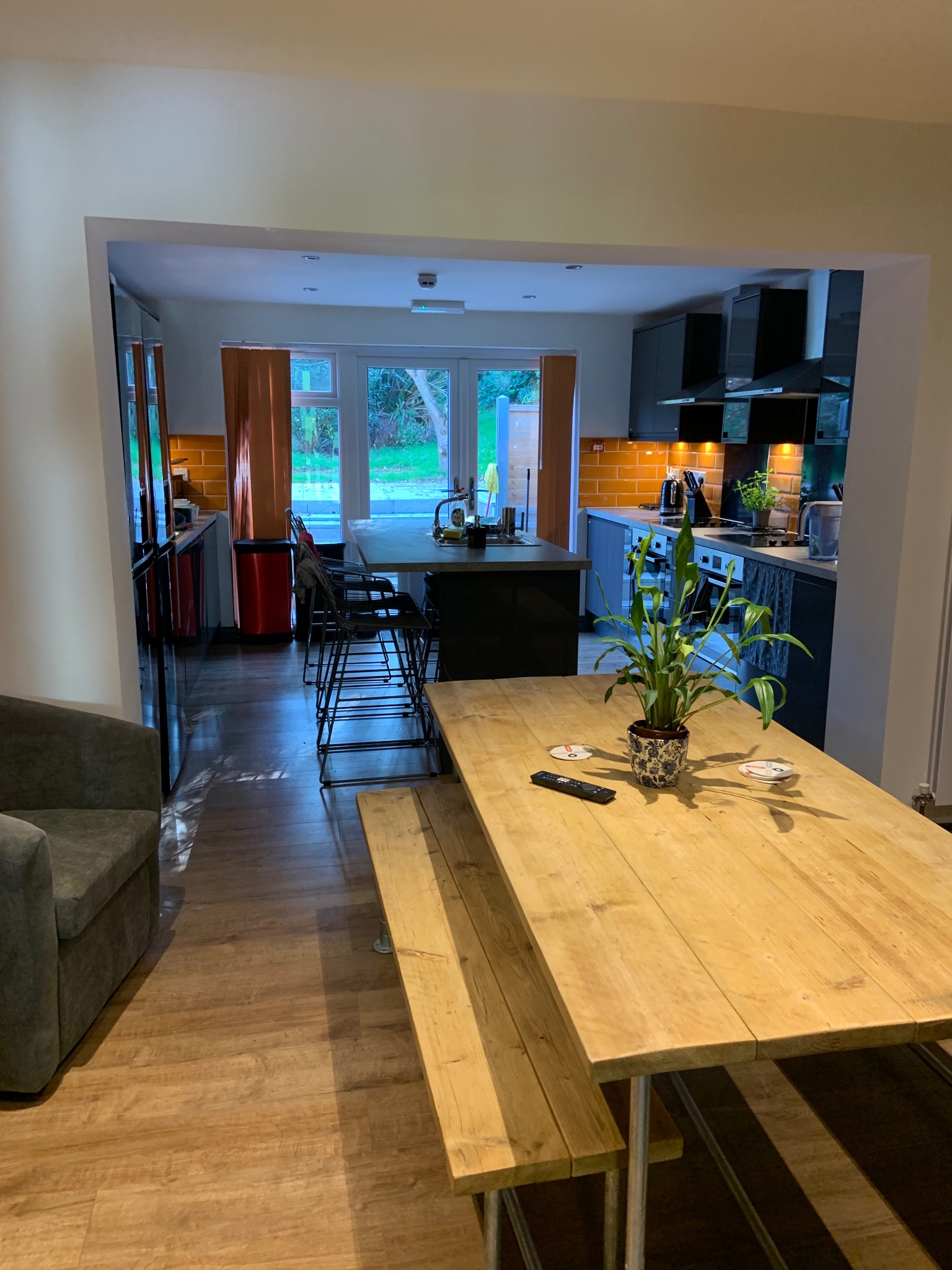 Nowoczesna kuchnia z drewnianym stołem i ławą. Pomarańczowe płytki na ścianie, czarne szafki i duży stół z rośliną w doniczce tworzą przytulną przestrzeń.