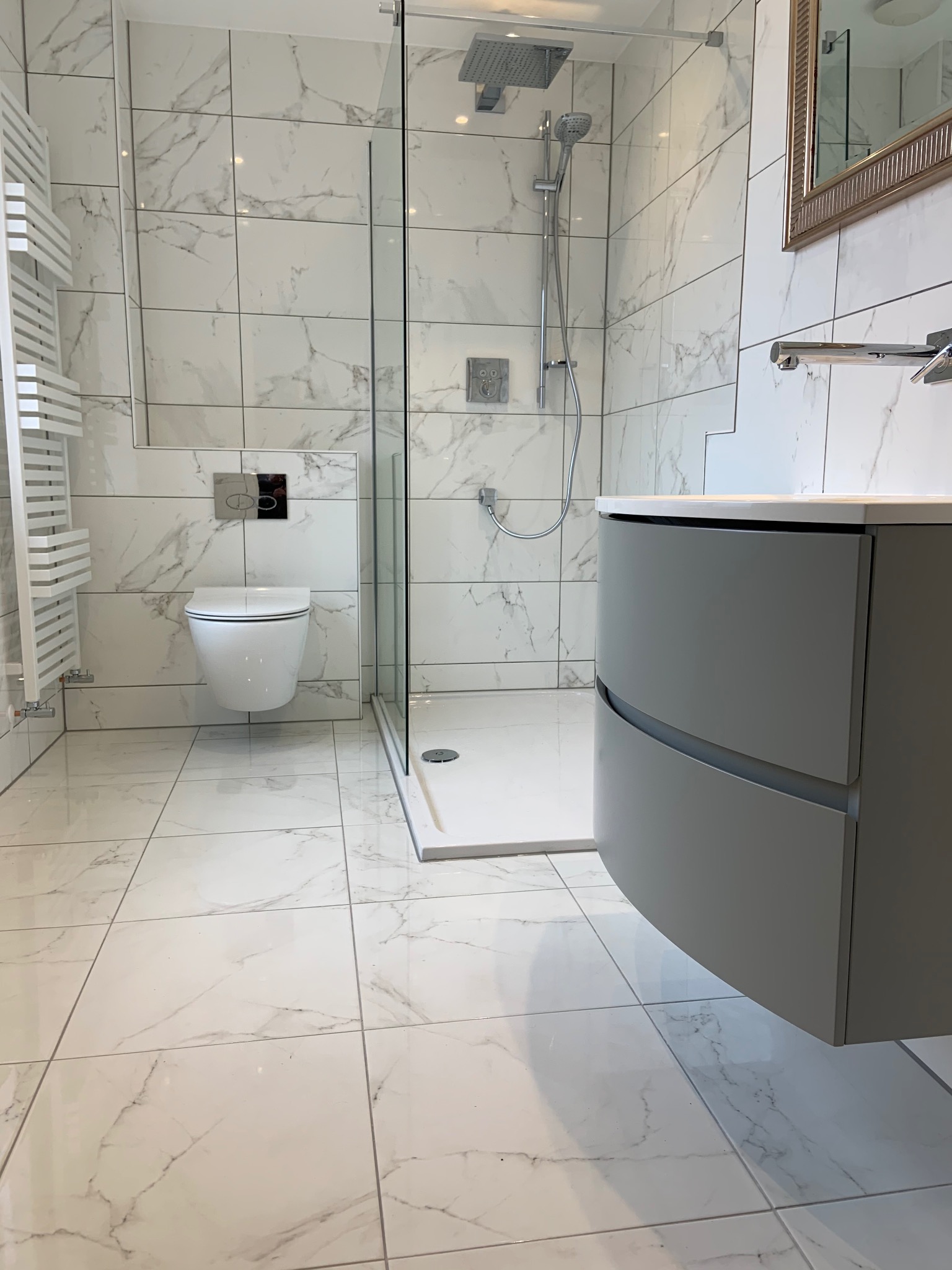 Nowoczesna łazienka z kabiną prysznicową typu walk-in, umywalką z szafką w odcieniach szarości i marmurowymi płytkami na ścianach i podłodze. Minimalistyczny grzejnik.