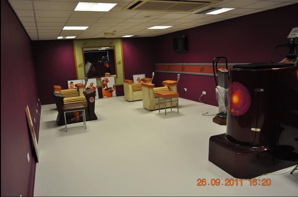 Nowoczesne wnętrze studia masażu z fioletowymi ścianami, jasną podłogą i specjalistycznymi urządzeniami do masażu. Widoczne plakaty reklamowe i oświetlenie sufitowe.