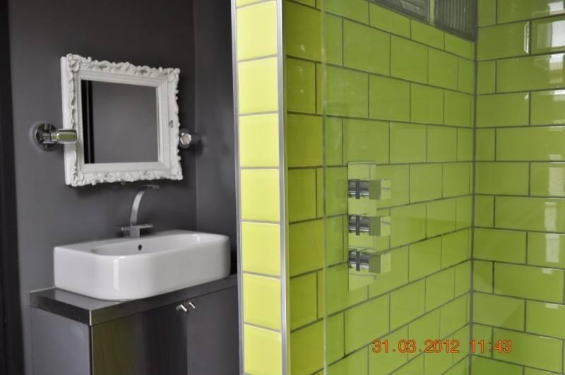 Nowoczesna łazienka z zielonymi płytkami, białą umywalką i ozdobnym lustrem w białej ramie. Szara ściana i chromowana armatura. Widoczny fragment kabiny prysznicowej.