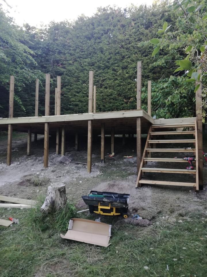 Budowa drewnianego tarasu na nierównym terenie, widoczne słupy, podest i schody. W tle drzewa, na pierwszym planie narzędzia i resztki materiałów budowlanych.