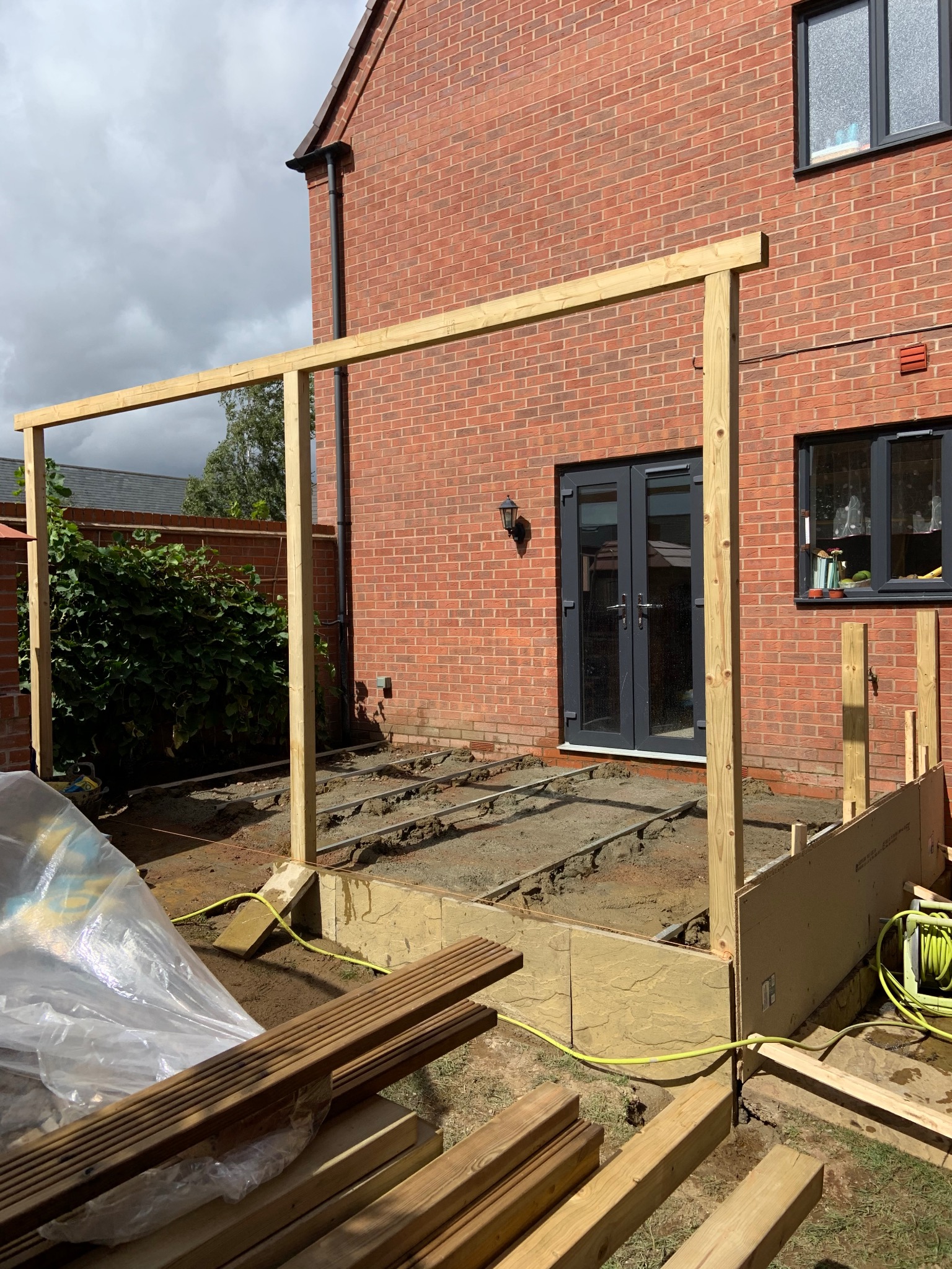 Konstrukcja drewniana tarasu w trakcie budowy przy domu z cegły. Widoczne deski i elementy montażowe. Etap przygotowania podłoża i wznoszenia ramy nośnej.