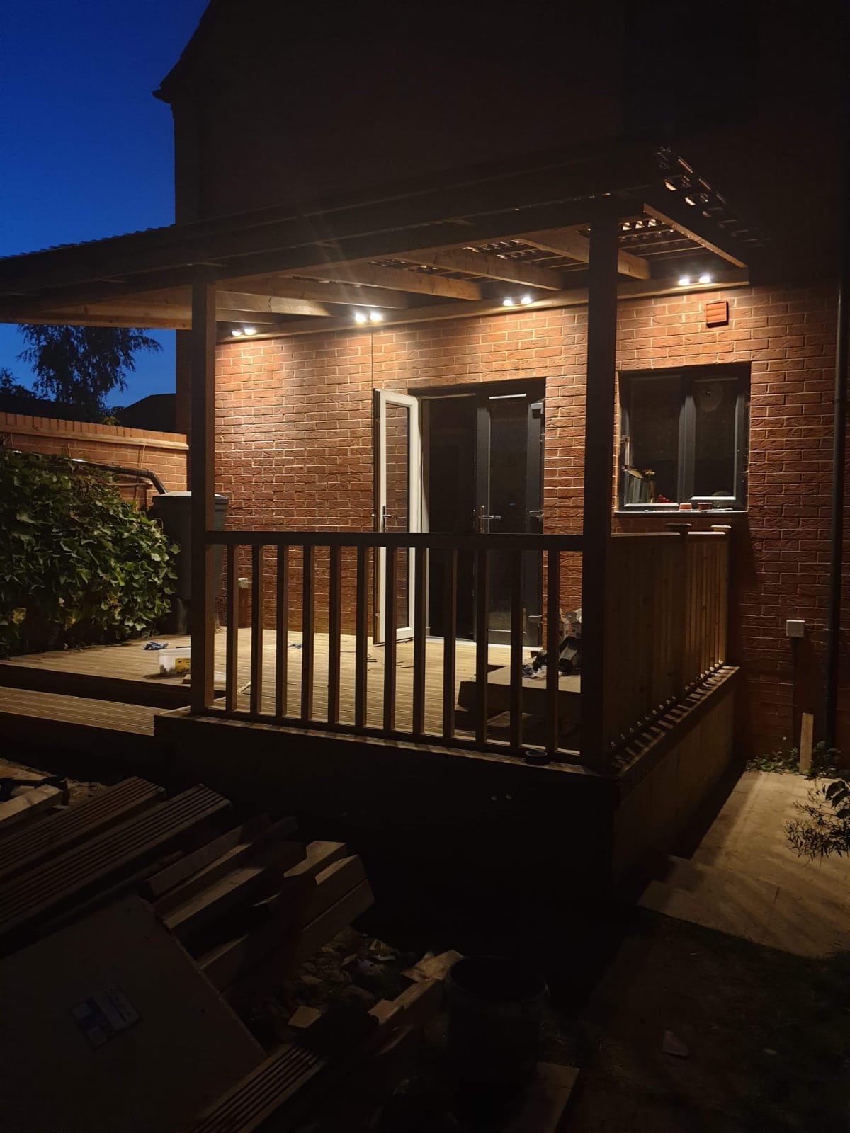 Zadaszony taras z drewnianą balustradą i oświetleniem punktowym przy ceglanym domu, wykonany w Łodzi. Widok nocny, częściowo widoczne materiały budowlane.