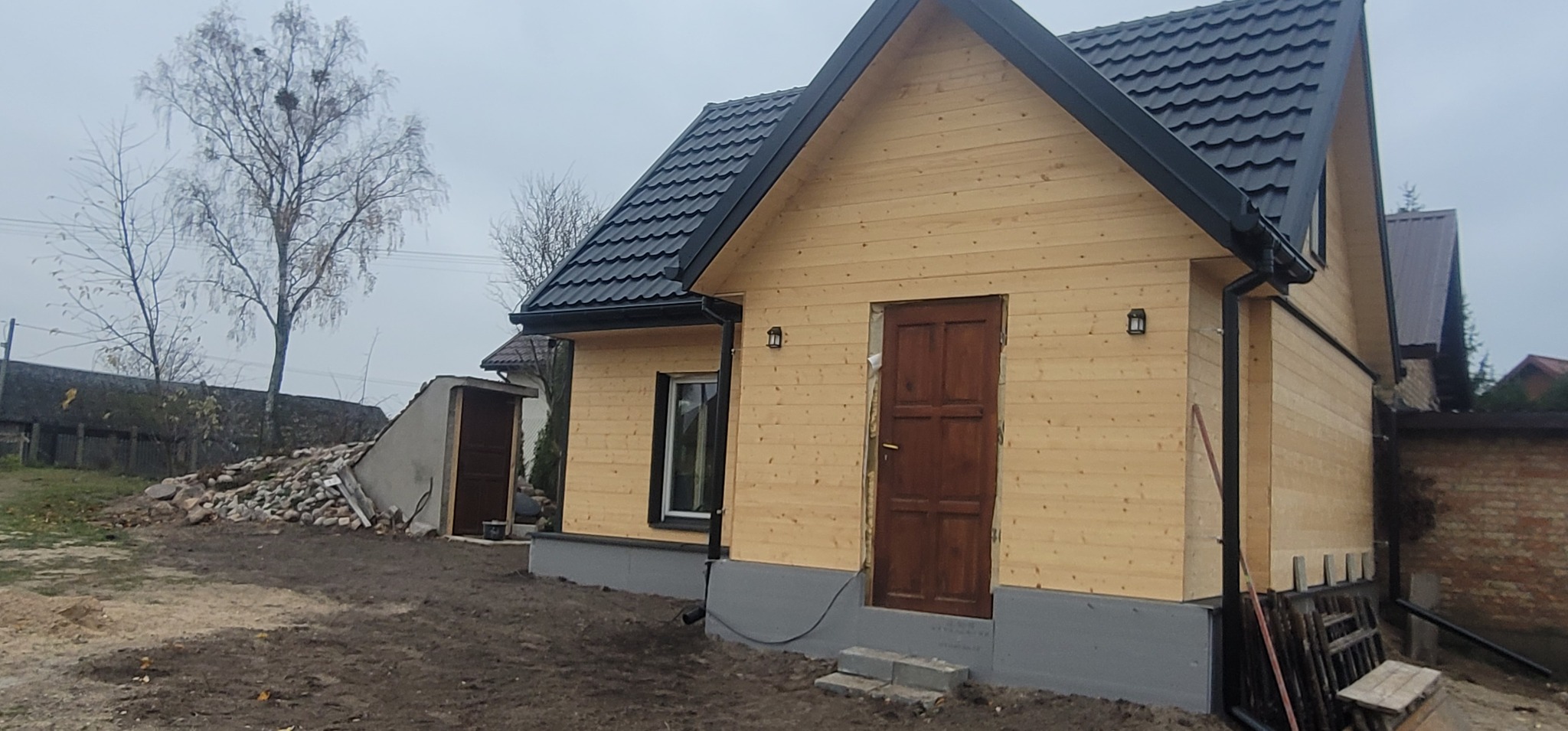 Drewniany dom z czarnym dachem i rynnami, oświetlony lampami zewnętrznymi, z widocznymi elementami wykończenia elewacji i otoczenia.