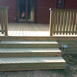 Karol Demko - Drewniany taras z balustradą i schodami, świeżo po budowie, widoczne gniazdka elektryczne na elewacji. W tle dom z bali i osoba w drzwiach.