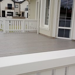 Karol Demko - Szara, drewniana podłoga tarasu z białymi balustradami. Widoczne fragmenty elewacji budynku i okna. Na ścianie wystające kable. Nowoczesne budownictwo.