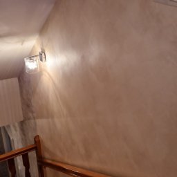 Karol Demko - Ściana w odcieniach beżu z dekoracyjną fakturą, oświetlona kinkietem. Widoczne drewniane schody i balustrada. Detal wykończenia wnętrza.