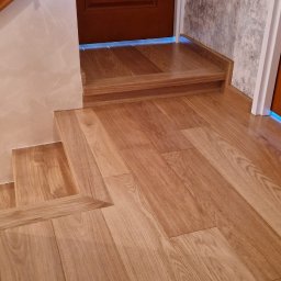 Karol Demko - Drewniane schody i podłoga z jasnego parkietu, brązowe drzwi z napisem 'bad boy' w tle, fragment balustrady po lewej stronie. Detal wykończenia wnętrza.