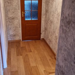 Karol Demko - Korytarz po remoncie: drewniana podłoga, ściany z efektem betonu, brązowe drzwi z szybą. Widoczne listwy przypodłogowe i oświetlenie sufitowe.