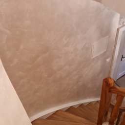 Karol Demko - Spiralne, drewniane schody w jasnym wnętrzu z dekoracyjną ścianą w odcieniach beżu. Widoczne drewniane poręcze i fragment drzwi.