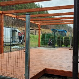 Karol Demko - Drewniana pergola z metalowymi słupkami, świeżo polakierowany taras. W tle las, krzewy i bus z otwartymi drzwiami. Człowiek kuca za ogrodzeniem.