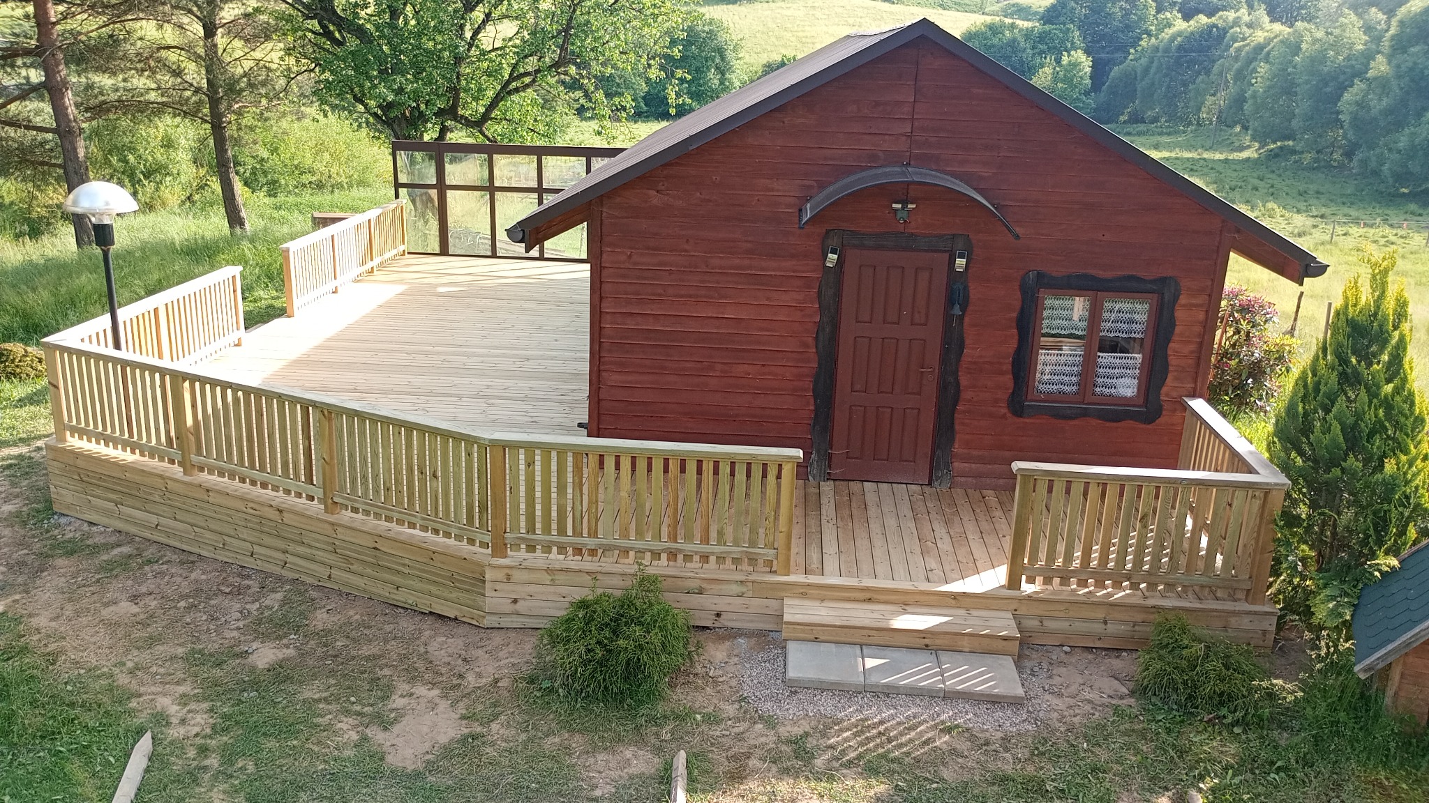 Drewniany domek z tarasem i balustradą, otoczony zielenią. Widok z góry ukazuje detale konstrukcji i naturalne otoczenie.