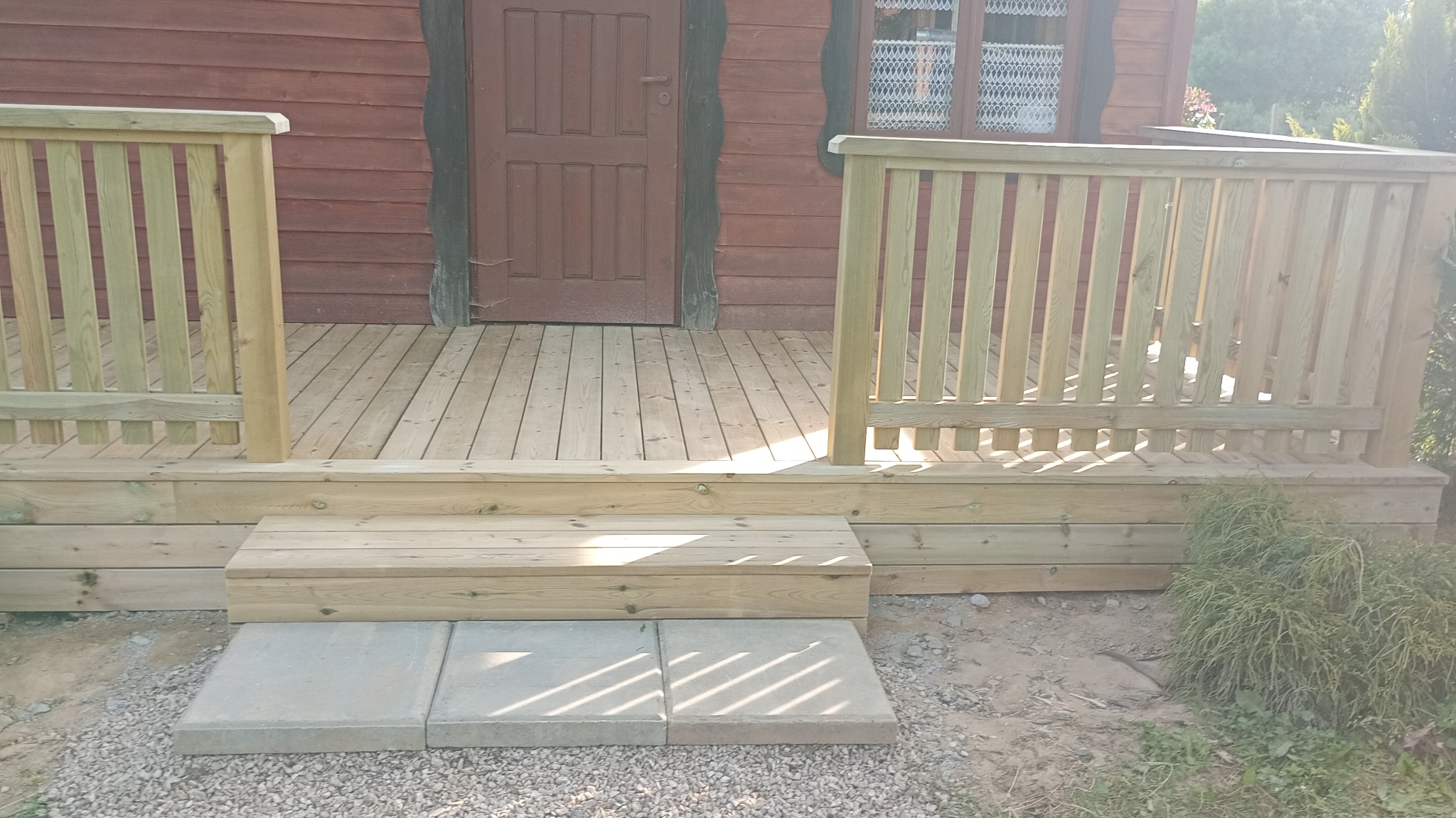 Drewniany ganek z balustradą przy brązowym domu z oknem. Schody z płyt betonowych. Naturalne światło podkreśla strukturę drewna.