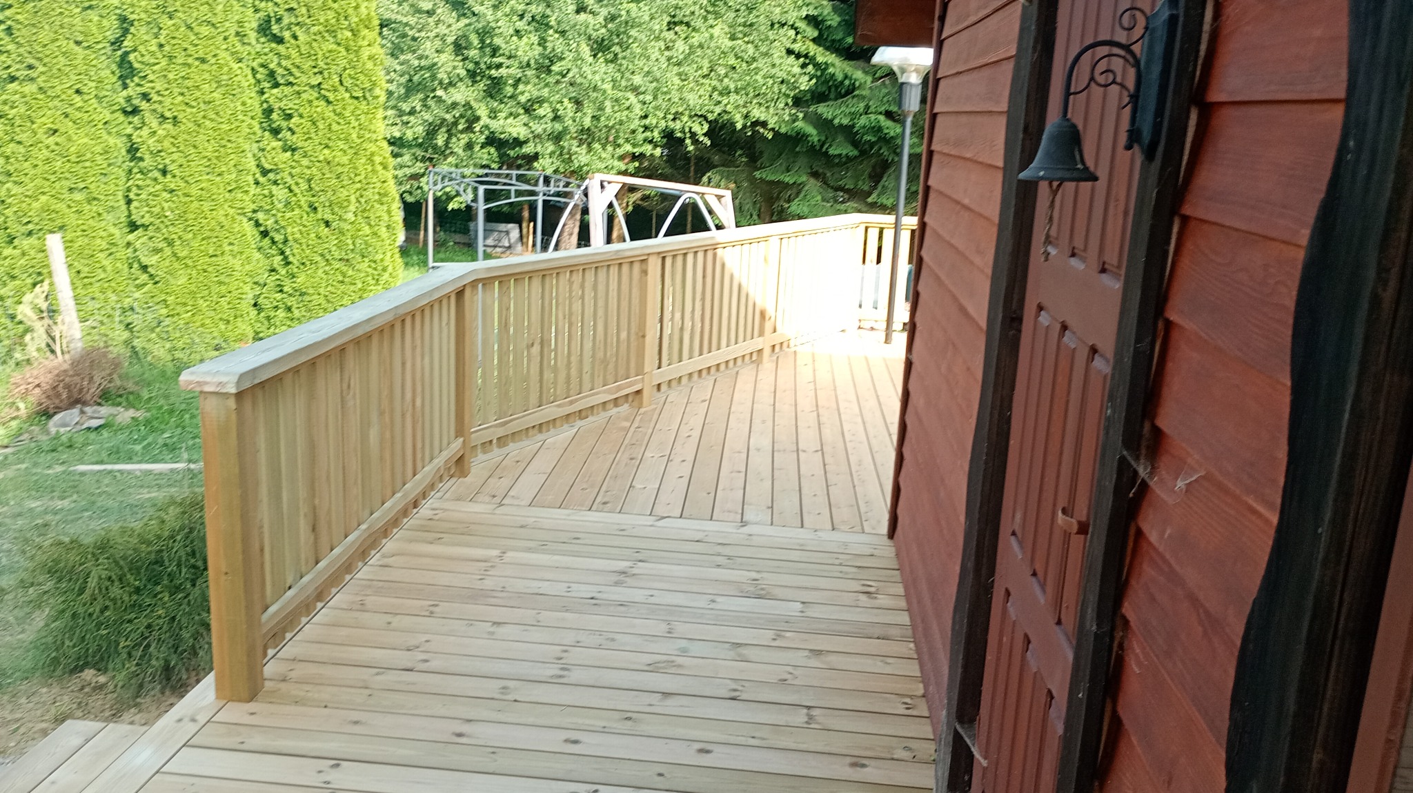 Nowa drewniana weranda z balustradą, z widokiem na zielony ogród i częściowo widoczną pergolę w tle. Ciemne drewno elewacji budynku.