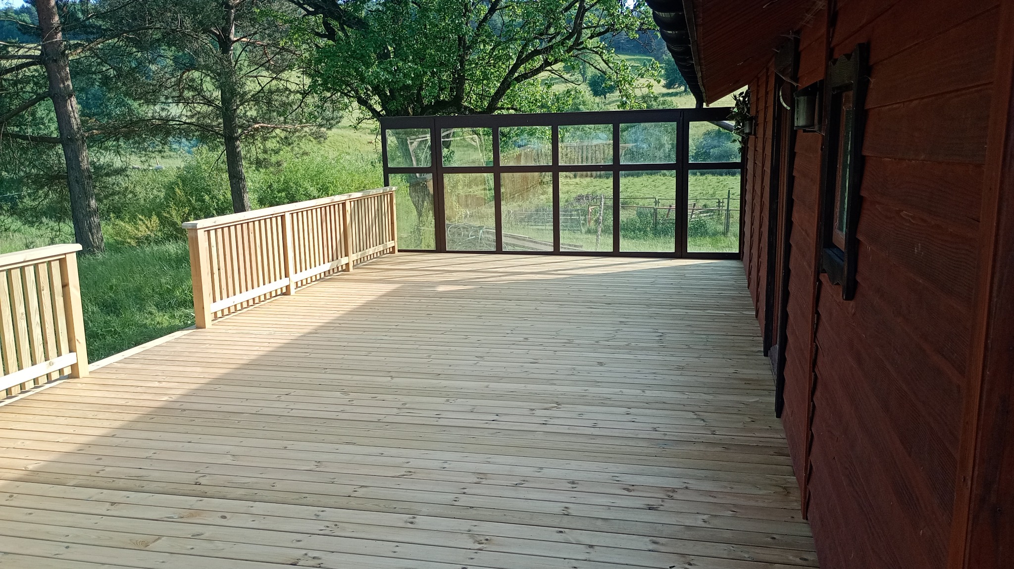 Nowy, jasny, drewniany taras z balustradą i przeszkloną ścianą, przylegający do budynku z ciemnego drewna, na tle zieleni drzew i łąki. Słoneczny dzień.