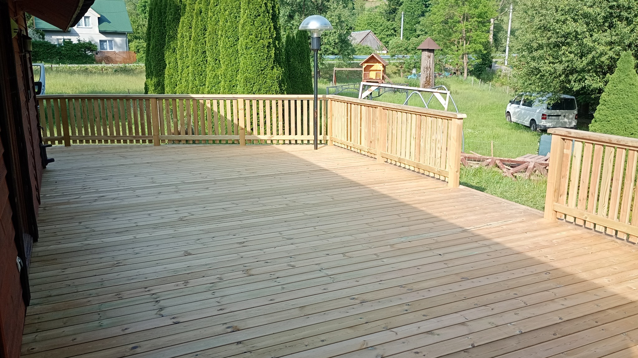 Drewniany taras z jasnymi deskami i balustradą, oświetlony lampą, z widokiem na zieleń i zabudowania w tle. Świeżo zaimpregnowane deski.