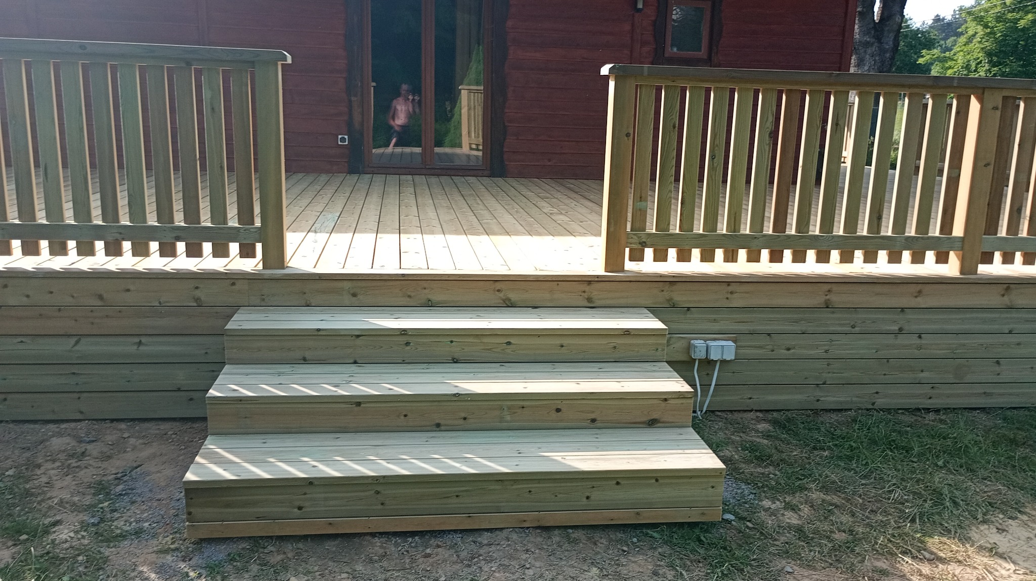 Drewniany taras z balustradą i schodami, świeżo po budowie, widoczne gniazdka elektryczne na elewacji. W tle dom z bali i osoba w drzwiach.