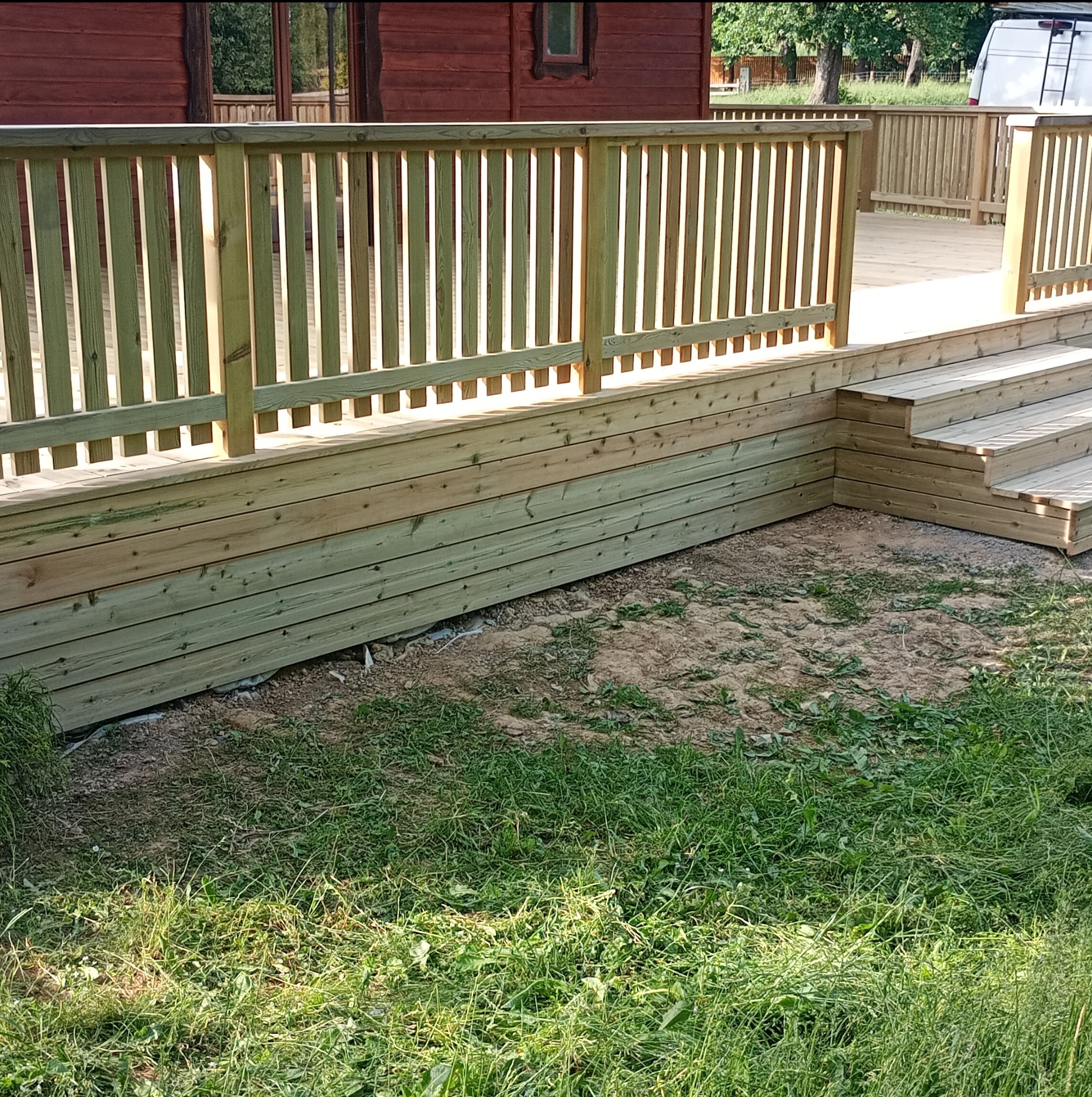 Nowa drewniana konstrukcja tarasu z balustradą i schodami, świeżo po impregnacji, na tle trawnika i budynku z czerwonego drewna.