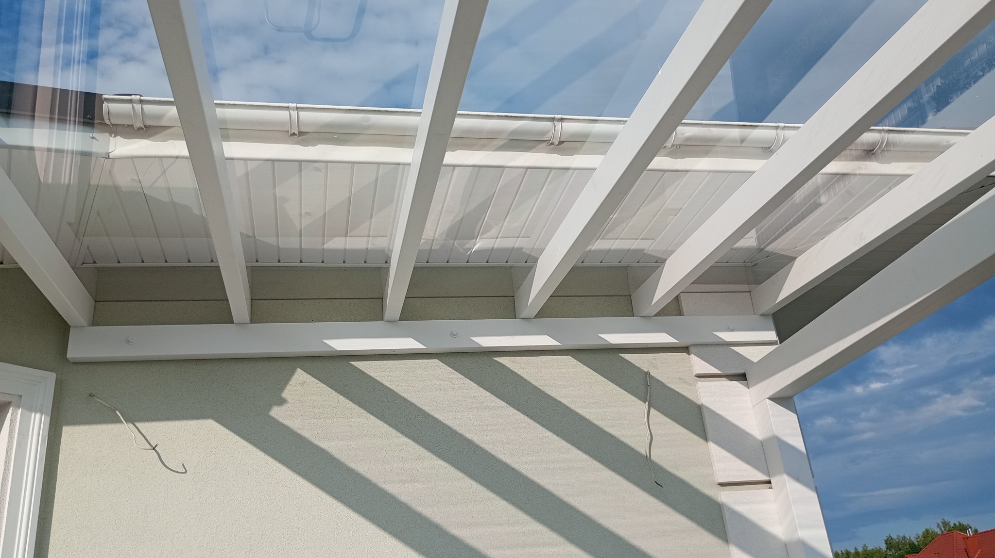 Biała pergola z przezroczystym zadaszeniem, widok z dołu, ukazujący konstrukcję i światło słoneczne. Elementy drewniane pomalowane na biało.