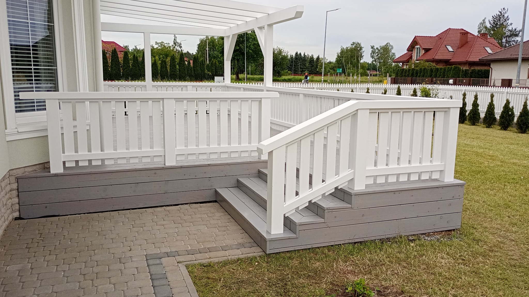 Biała pergola z balustradą i schodami z szarego drewna, widok z boku. W tle dom z czerwoną dachówką i zielony trawnik. Jasne, naturalne oświetlenie.