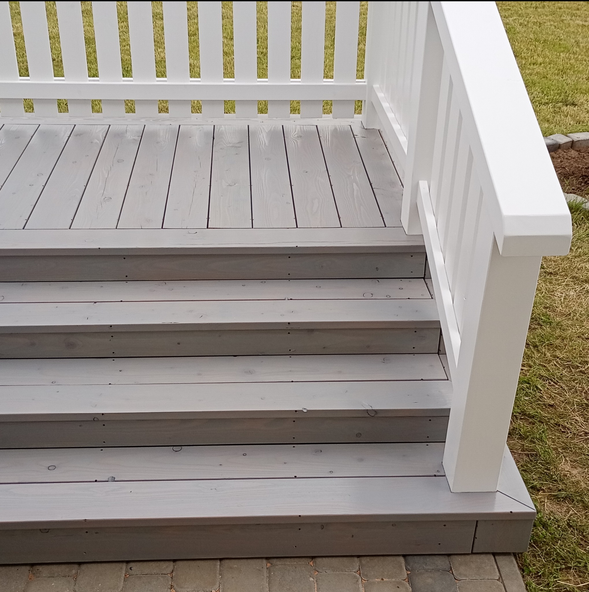 Schody drewniane i balustrada w jasnoszarym odcieniu. Widoczne ślady po wkrętach, świeżo pomalowane. Trawa w tle.