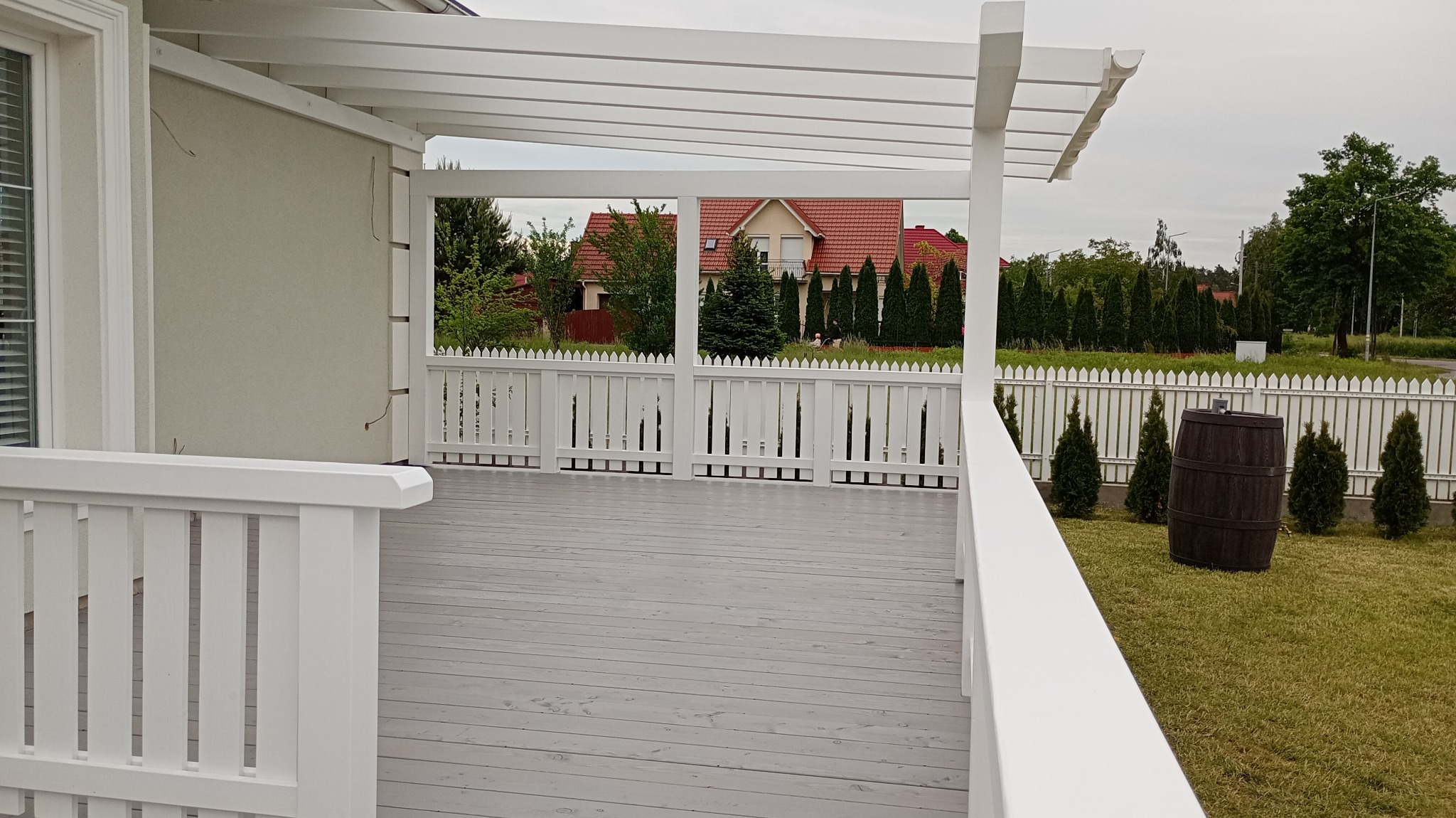Biała pergola z drewnianym, szarym podestem, widok na ogród z tujami i drewnianym beczką. W tle dom z czerwonym dachem.