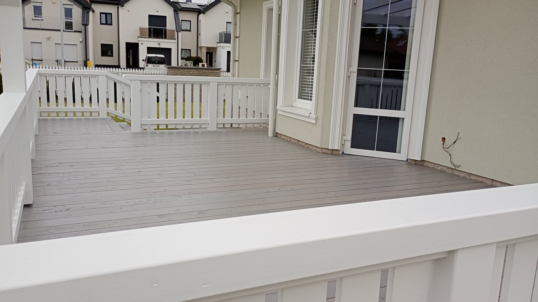 Szara, drewniana podłoga tarasu z białymi balustradami. Widoczne fragmenty elewacji budynku i okna. Na ścianie wystające kable. Nowoczesne budownictwo.