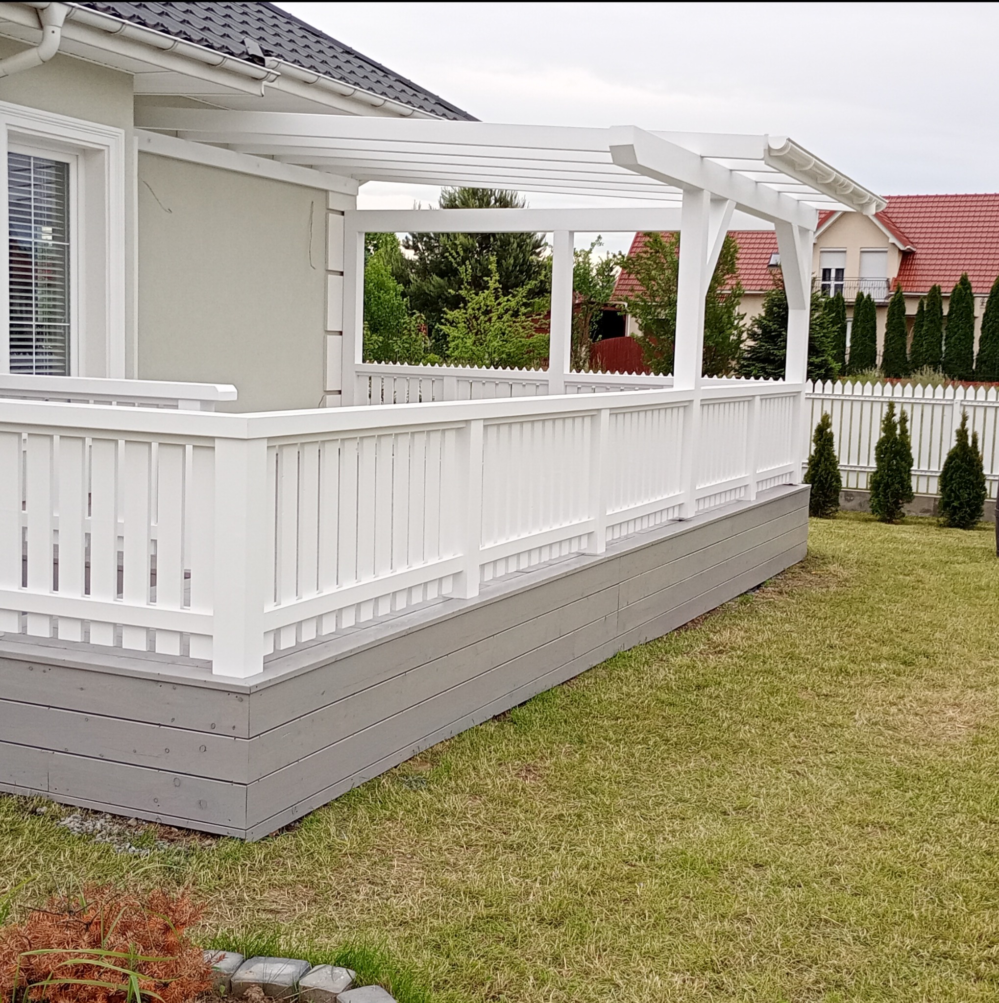 Biała pergola z balustradą, szara podstawa, widok na dom z czerwonym dachem i zielony trawnik. Klasyczny styl, staranne wykonanie.