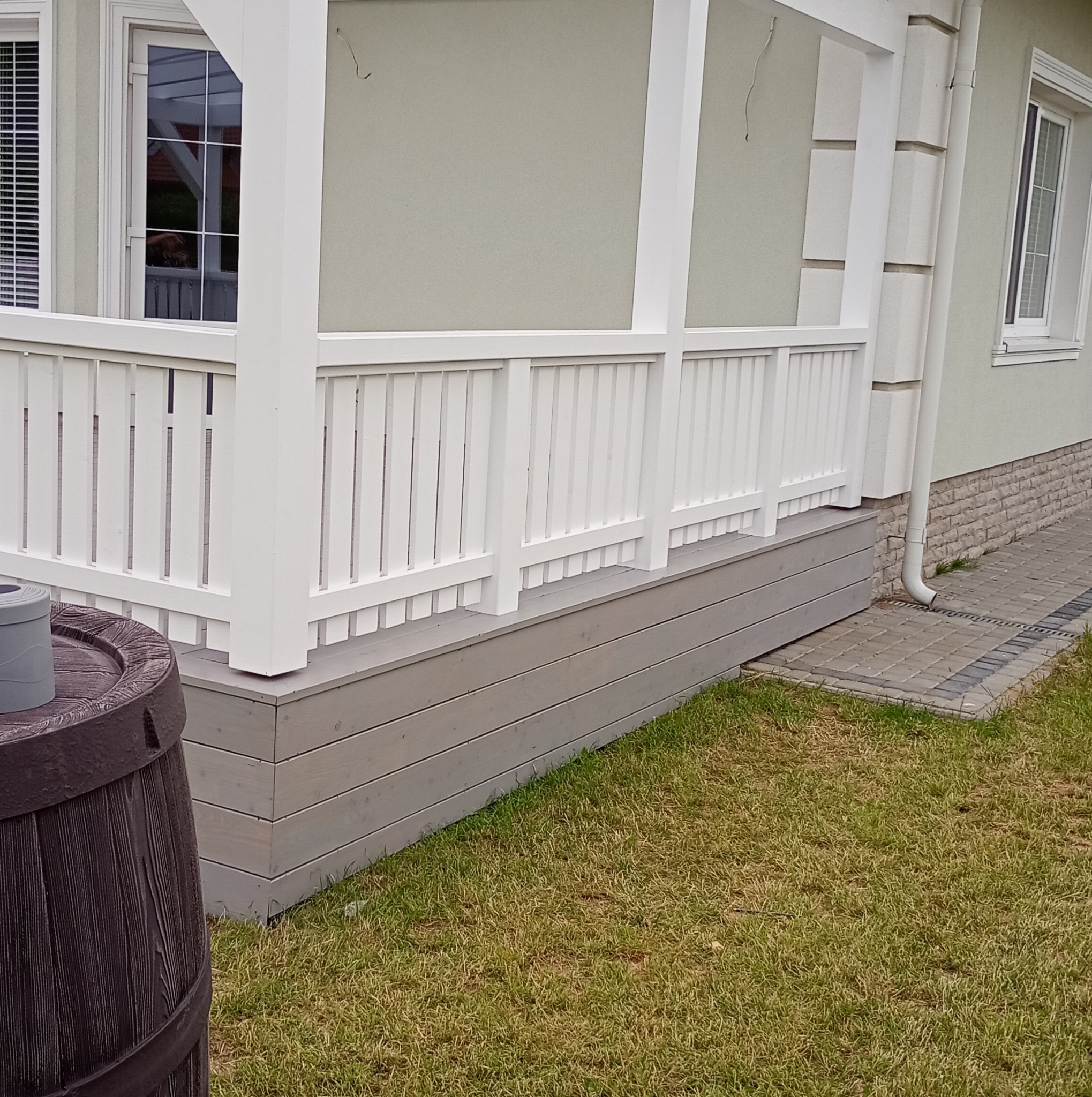 Biała, drewniana balustrada tarasu przydomowego z szarym podestem, widok z poziomu trawnika, z fragmentem dekoracyjnej beczki na pierwszym planie.