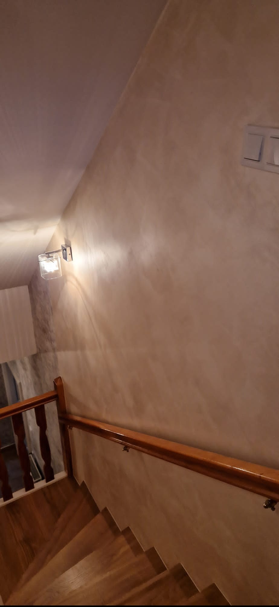 Ściana w odcieniach beżu z dekoracyjną fakturą, oświetlona kinkietem. Widoczne drewniane schody i balustrada. Detal wykończenia wnętrza.