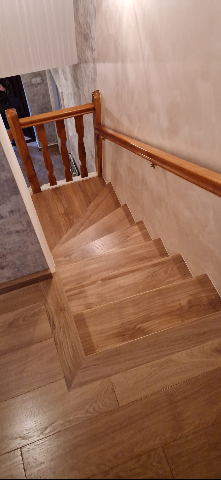 Drewniane schody z balustradą w jasnym odcieniu, prowadzące w dół. Ściany w beżowym odcieniu, podłoga wyłożona parkietem. Widok z góry.