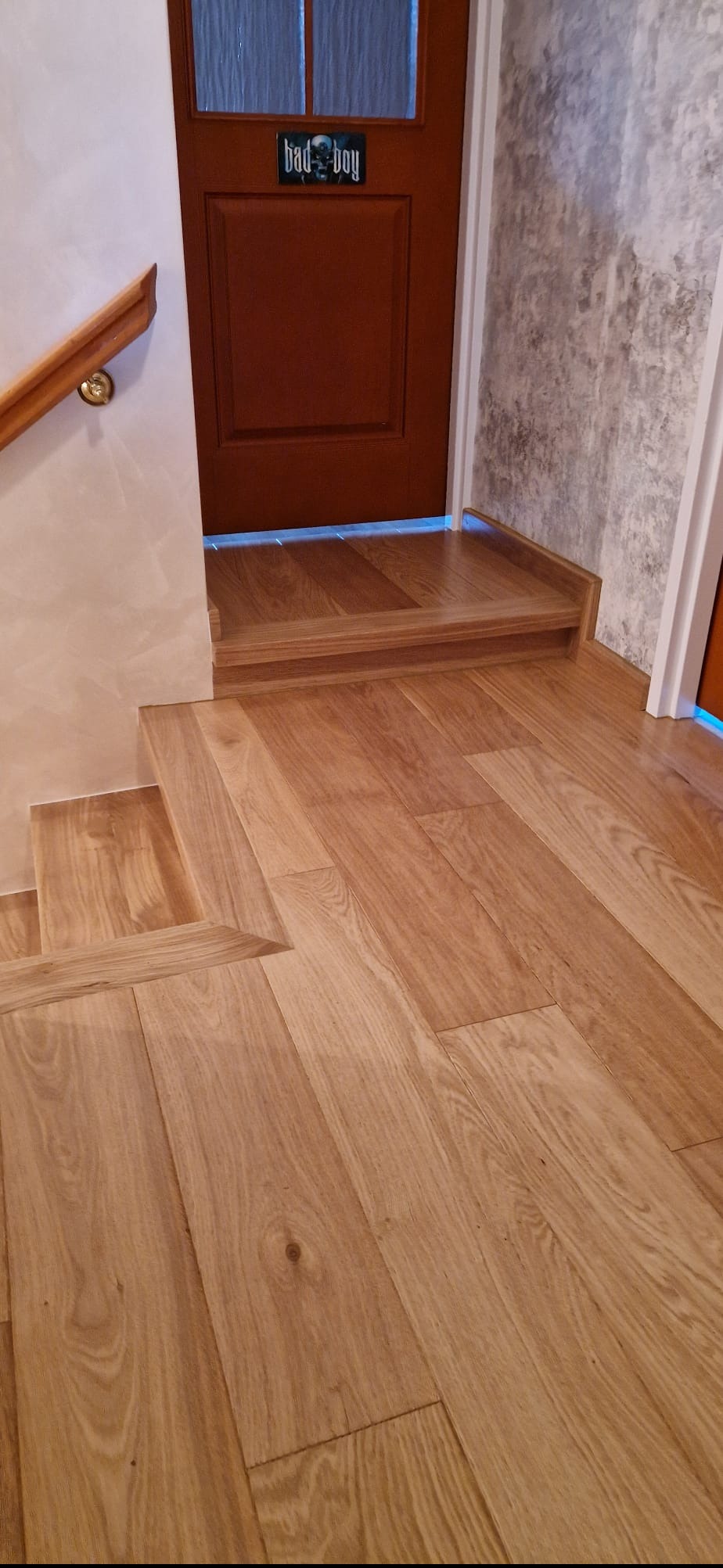 Drewniane schody i podłoga z jasnego parkietu, brązowe drzwi z napisem 'bad boy' w tle, fragment balustrady po lewej stronie. Detal wykończenia wnętrza.