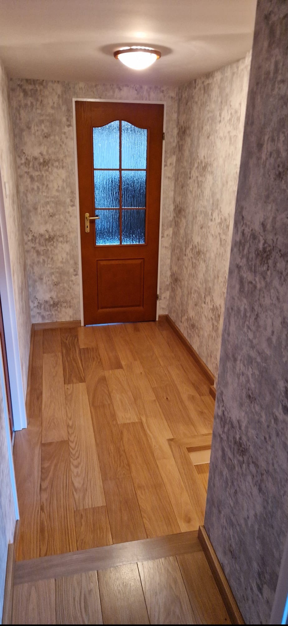 Korytarz po remoncie: drewniana podłoga, ściany z efektem betonu, brązowe drzwi z szybą. Widoczne listwy przypodłogowe i oświetlenie sufitowe.