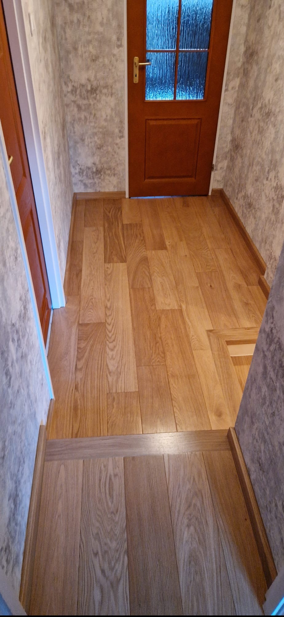 Ukończony korytarz z nową, jasną podłogą drewnianą i odnowionymi ścianami o szarym wzorze. Widoczna brązowa stolarka drzwiowa z przeszkleniem.