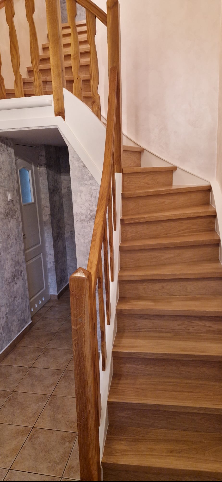 Drewniane schody z balustradą, widok z dołu. Jasne drewno, białe wykończenia. W tle widoczna ściana w odcieniach szarości i drzwi.