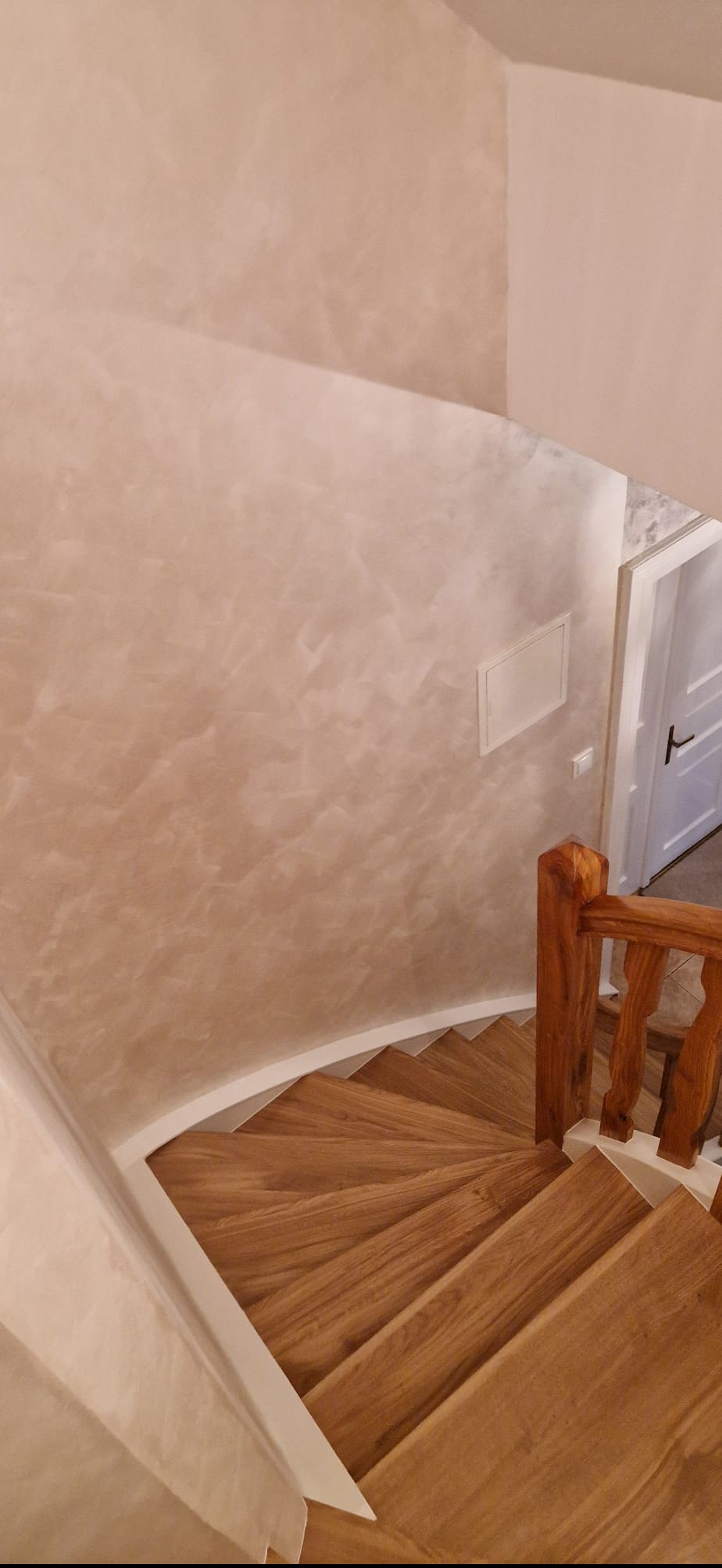 Klatka schodowa z drewnianymi schodami i ścianą o dekoracyjnym wykończeniu w odcieniach beżu. Widok z góry, podkreślający spiralny kształt schodów i unikalną fakturę ściany.