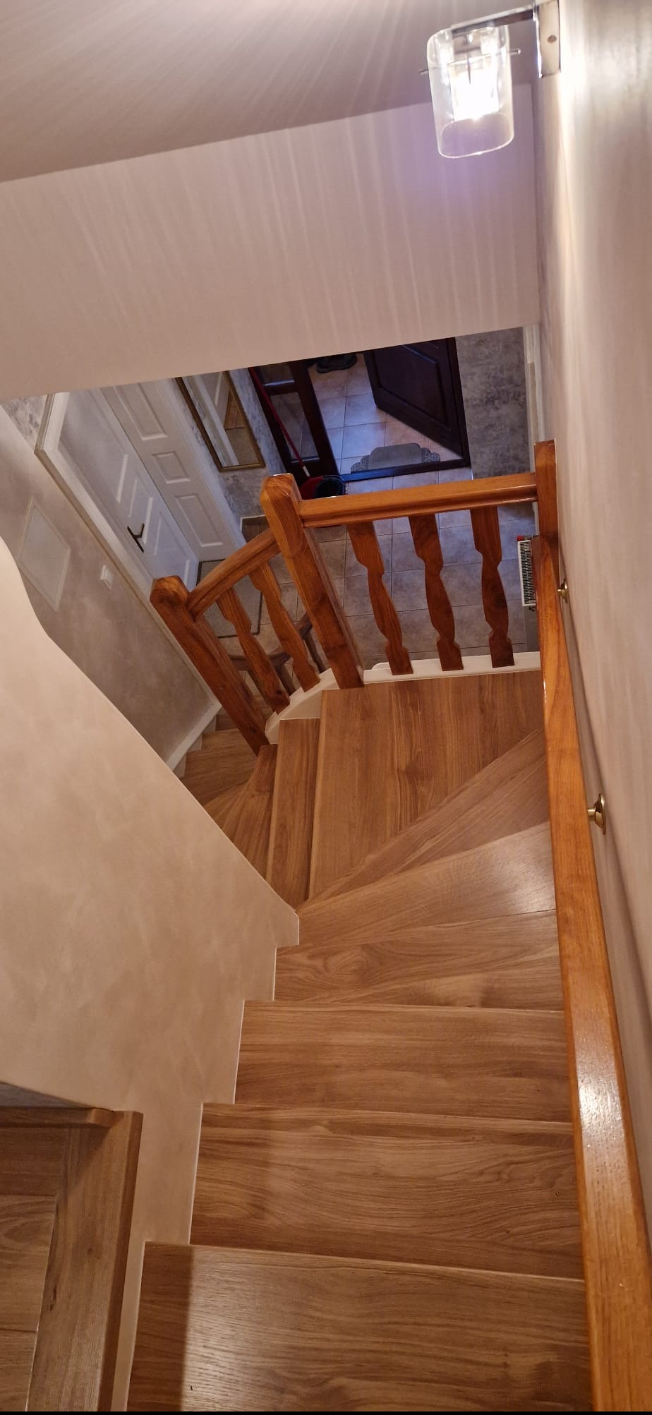 Drewniane schody z balustradą o ciepłym odcieniu, widok z góry, z oświetleniem sufitowym i fragmentem drzwi w tle. Wykończenie ścian w jasnym, neutralnym kolorze.