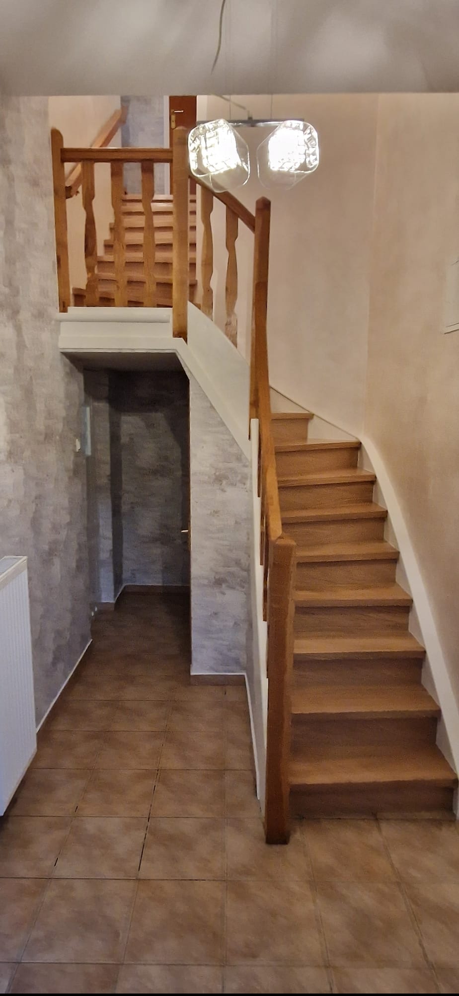 Jasne, drewniane schody z balustradą w dwukondygnacyjnym domu. Widoczne płytki podłogowe i dekoracyjna ściana. Nad schodami nowoczesna lampa.