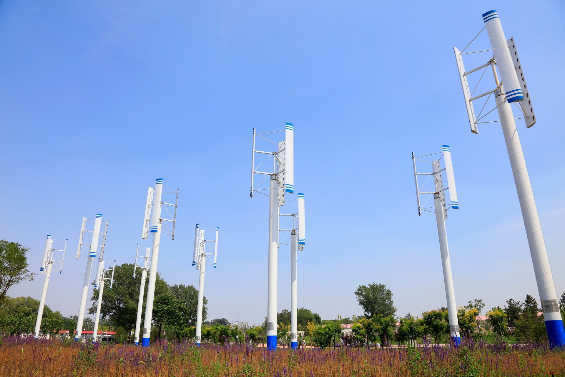 Białe turbiny wiatrowe pionowe z niebieskimi akcentami na tle błękitnego nieba i zieleni, widoczne z poziomu ziemi porośniętej fioletowymi kwiatami.