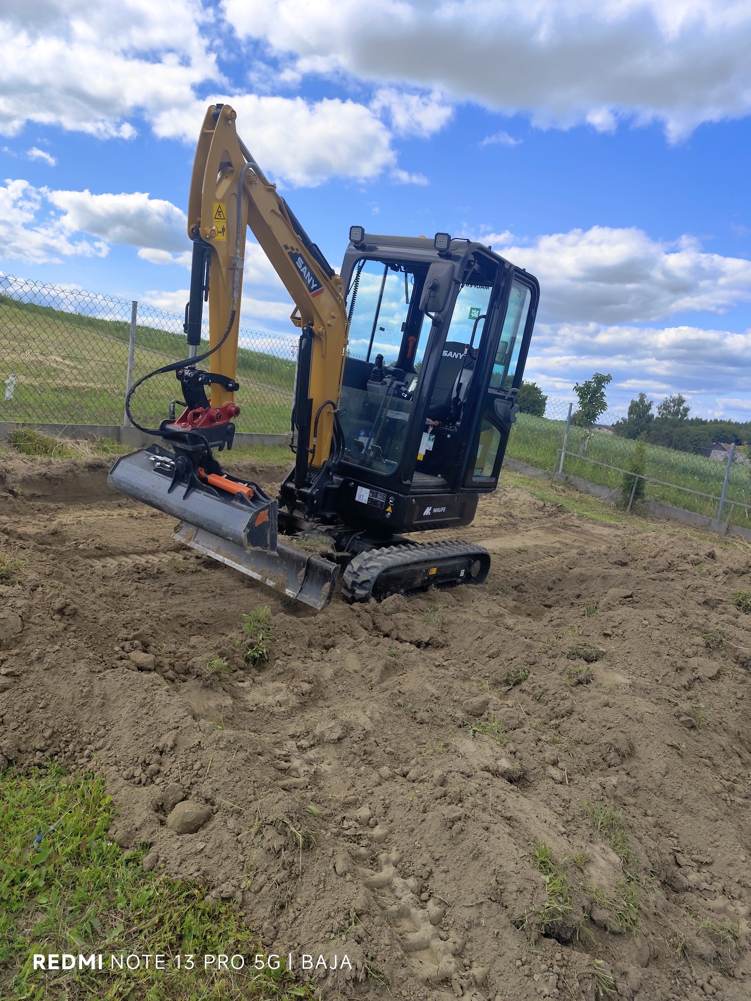 Żółto-czarna mini koparka SANY na nierównym terenie, z widocznym wykopem, pod błękitnym niebem z chmurami. Metalowe ogrodzenie w tle.