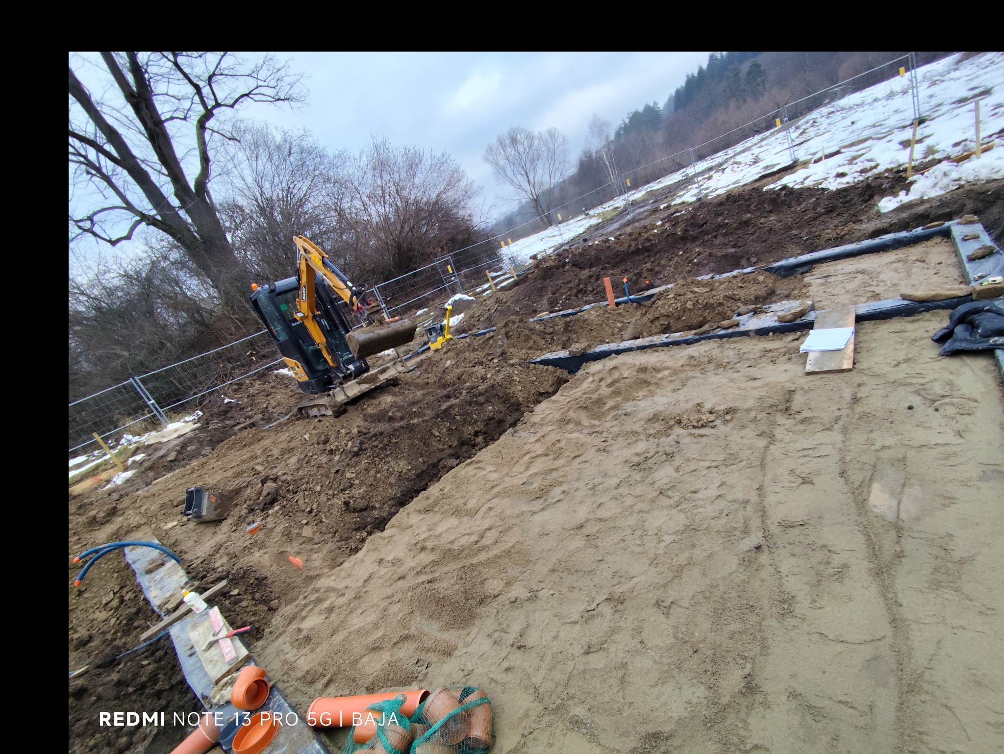 Żółta koparka wykonuje wykop pod fundamenty na zasypanym śniegiem terenie. Widoczne rury i zabezpieczenia wykopu. Ujęcie z boku, lekko pod kątem.