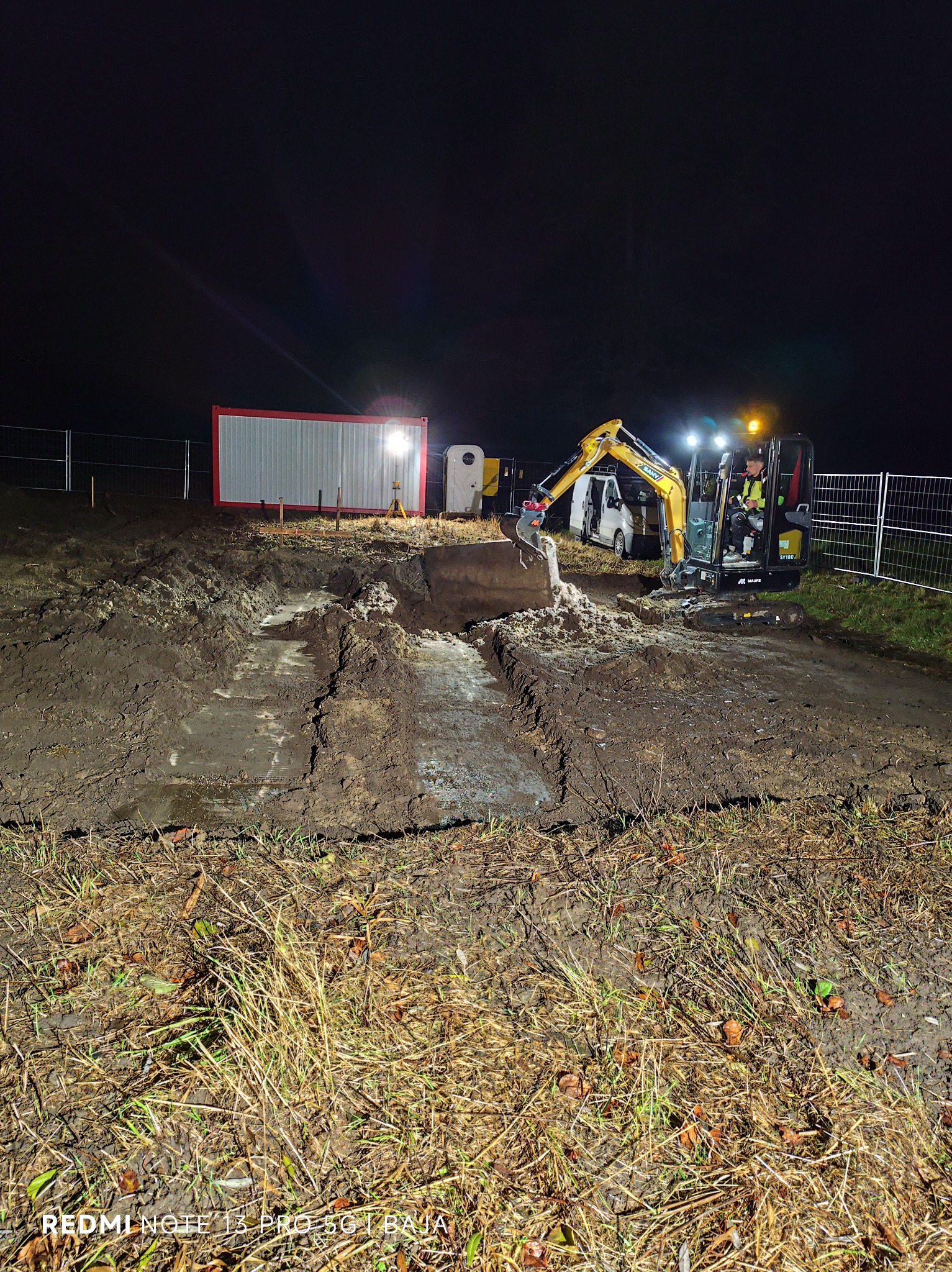 Żółta mini koparka SANY SY18C w akcji nocą, oświetlona reflektorami, wykonuje wykop w grząskim terenie. W tle kontener biurowy i biały van.
