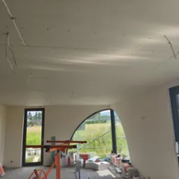Wnętrze w trakcie prac wykończeniowych: zamontowane płyty gipsowe na suficie, widoczne kable, worki z zaprawą i narzędzia. Nietypowe, łukowe okno z widokiem na zewnątrz.