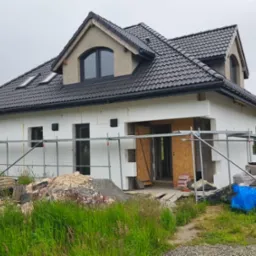 Dom w trakcie ocieplania styropianem, widoczne rusztowania i materiały budowlane. Dach pokryty ciemną dachówką, okna w szarych ramach. Trawa na pierwszym planie.