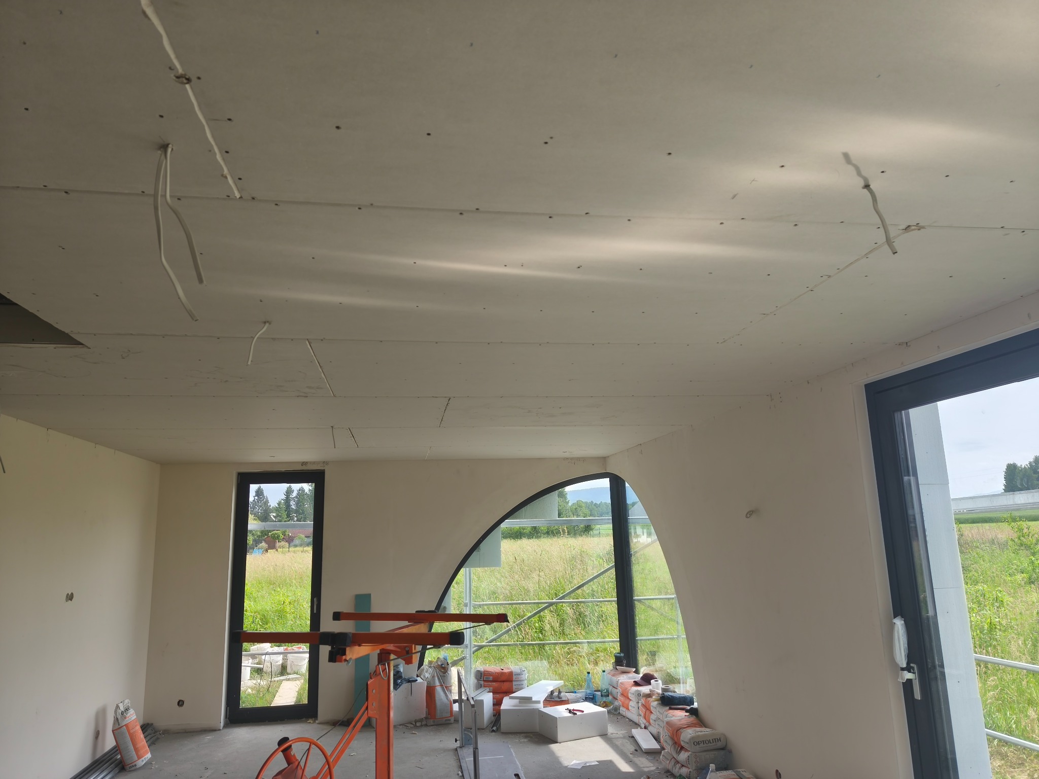 Wnętrze w trakcie prac wykończeniowych: zamontowane płyty gipsowe na suficie, widoczne kable, worki z zaprawą i narzędzia. Nietypowe, łukowe okno z widokiem na zewnątrz.