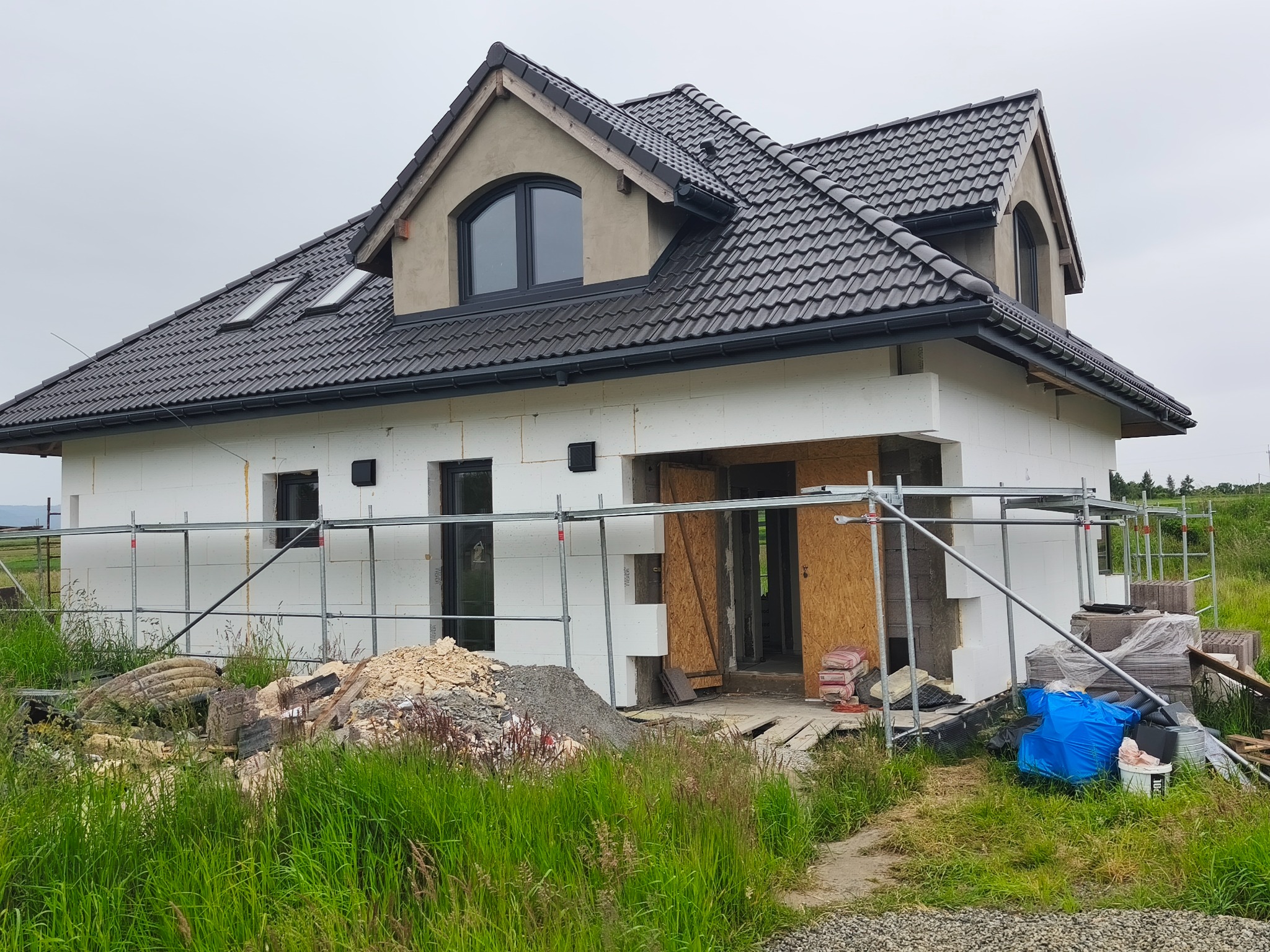 Dom w trakcie ocieplania styropianem, widoczne rusztowania i materiały budowlane. Dach pokryty ciemną dachówką, okna w szarych ramach. Trawa na pierwszym planie.