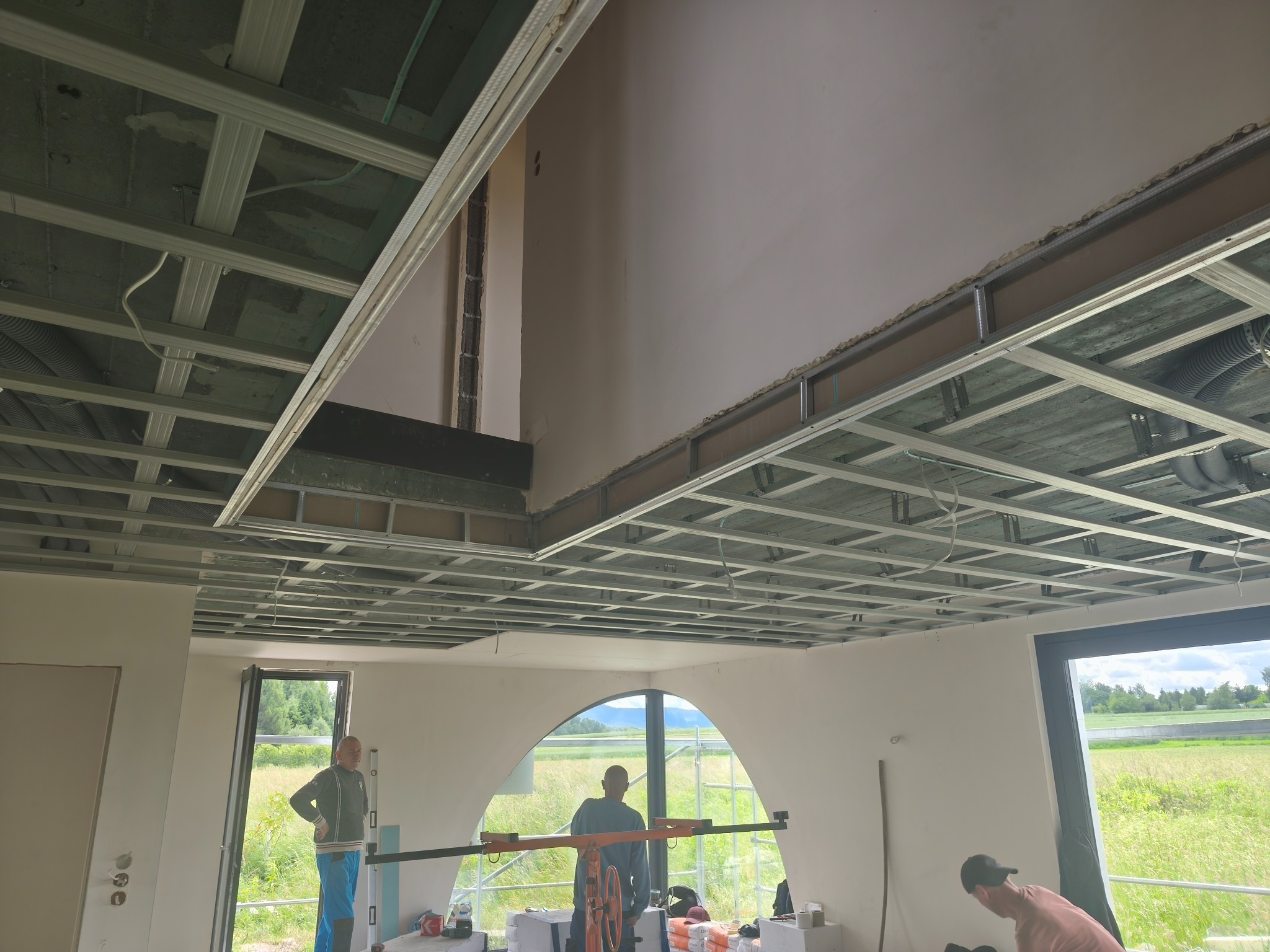 Montaż sufitu podwieszanego z metalowej konstrukcji w jasnym wnętrzu z dużymi oknami i widokiem na zewnątrz. Prace w toku, widoczni pracownicy i narzędzia.