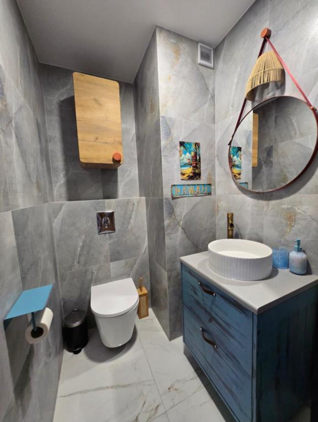 Nowoczesna łazienka z szarymi płytkami imitującymi marmur, drewnianą szafką wiszącą i okrągłym lustrem w skórzanej ramie. Biała umywalka na niebieskiej szafce.